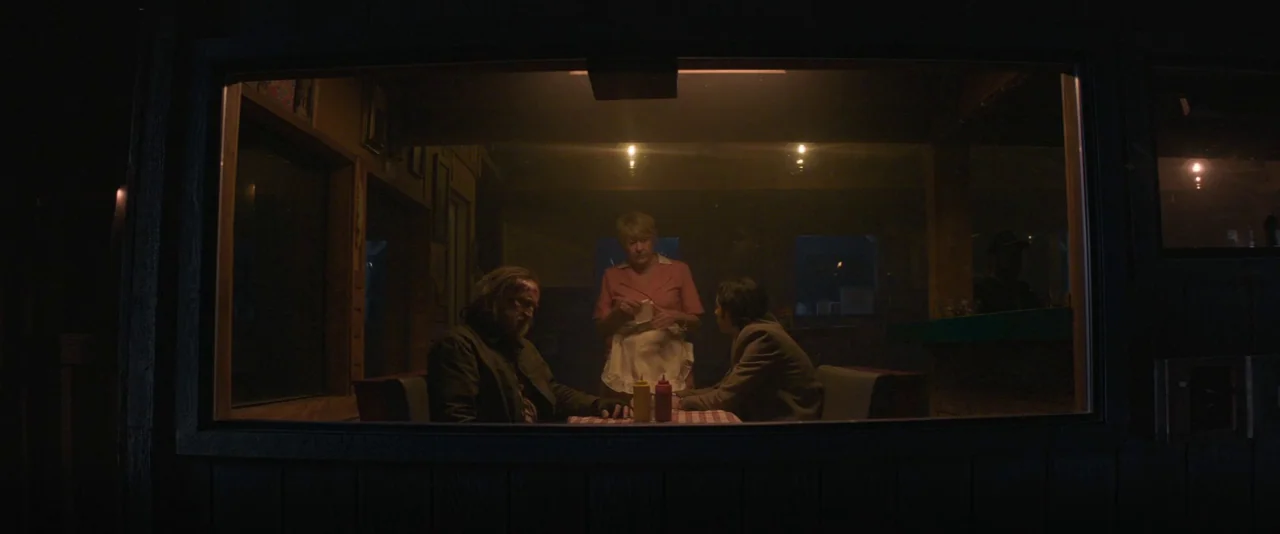 Pig (2021) movie still: wide — A dark diner booth features wooden walls and a checkered tablecloth illuminated from above. A middle-aged waitress…