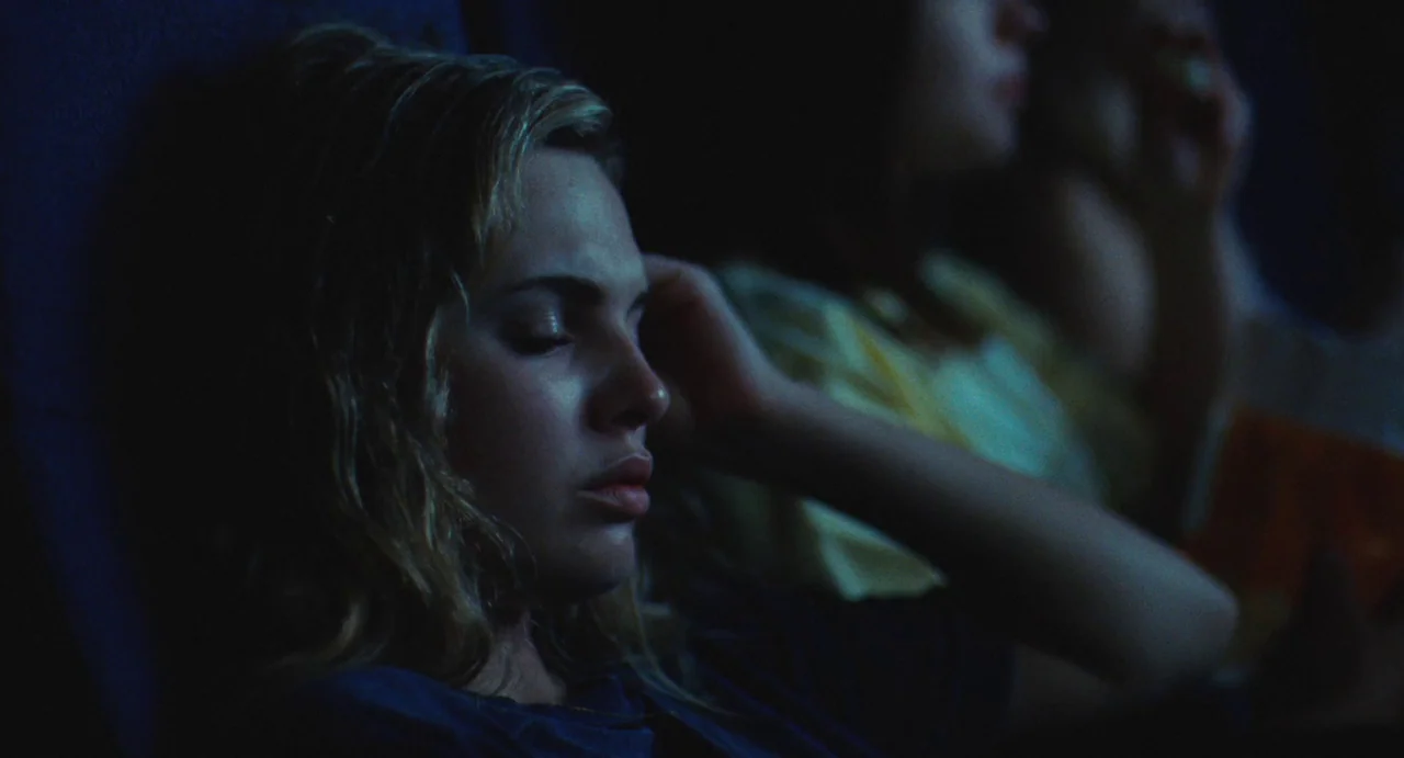 The Giant (2019) movie still: closeup — A dimly lit indoor auditorium with plush seats and dark surroundings. A young woman sits with her eyes…