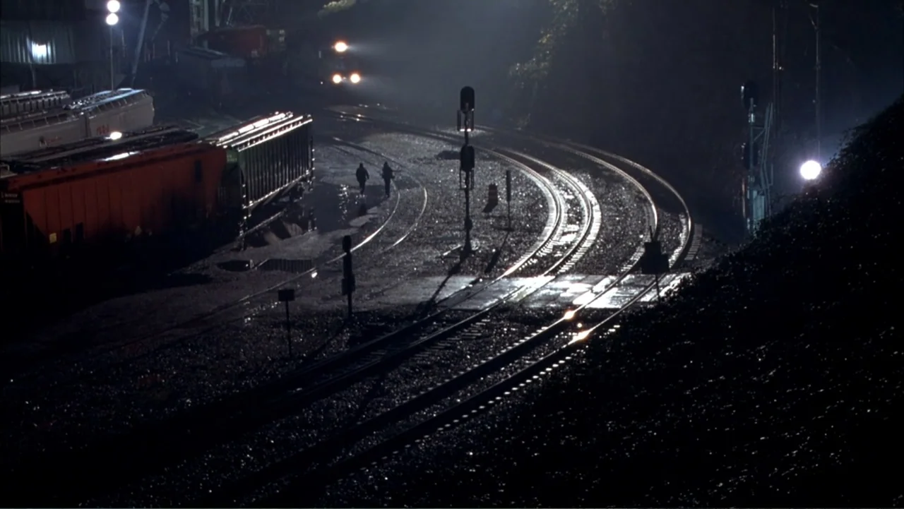 Paranoid Park (2007) movie still: wide — A sprawling nighttime industrial railroad yard features multiple tracks, train cars, and signal lights. Two…