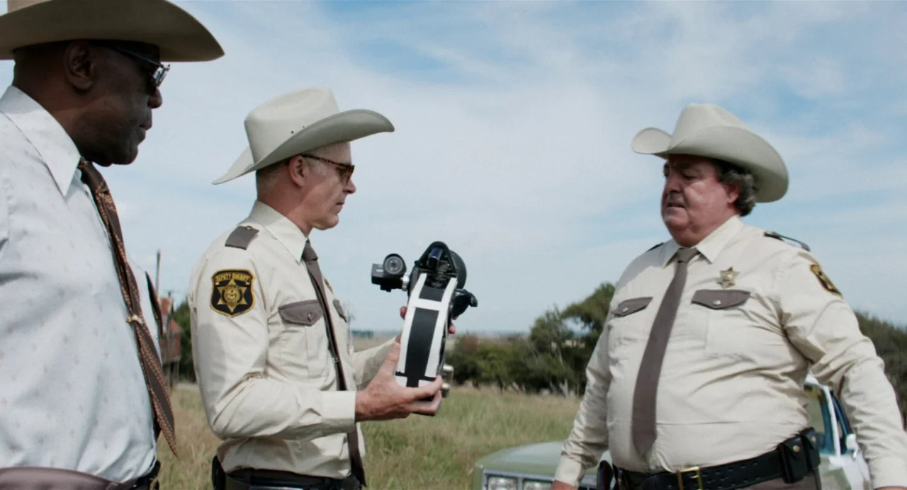 X (2022) movie still: medium — Three law enforcement officers stand in a grassy field on a bright day. One officer holds a technical device while…
