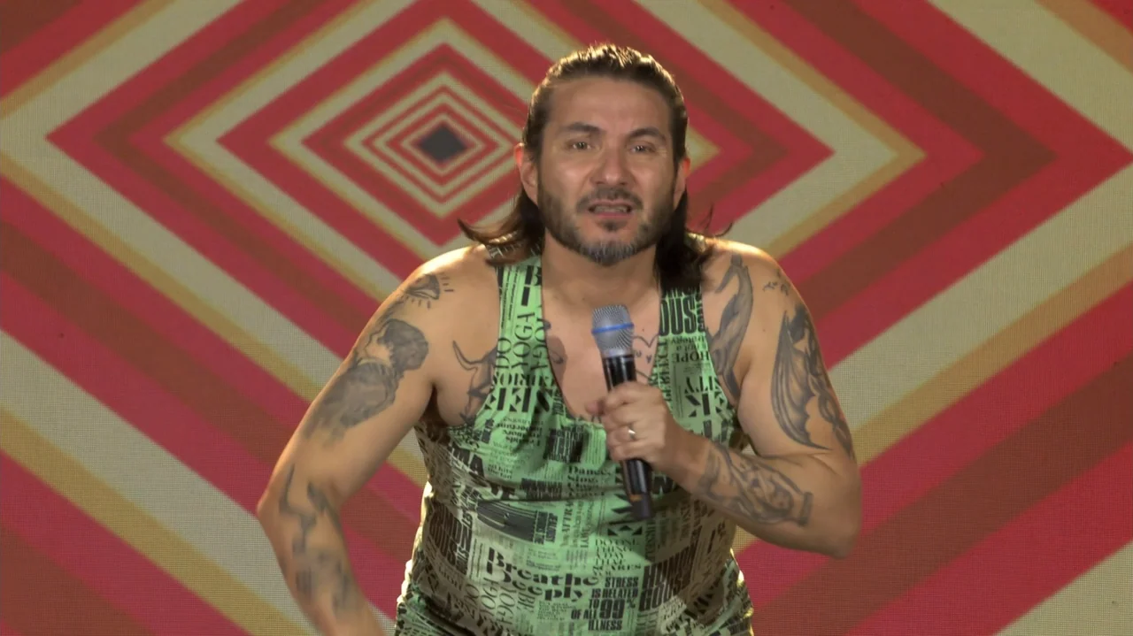 Felipe Avello: Doglover (2024) movie still: medium — A man stands on a stage in front of a red and white geometric wall. The man holds a microphone…