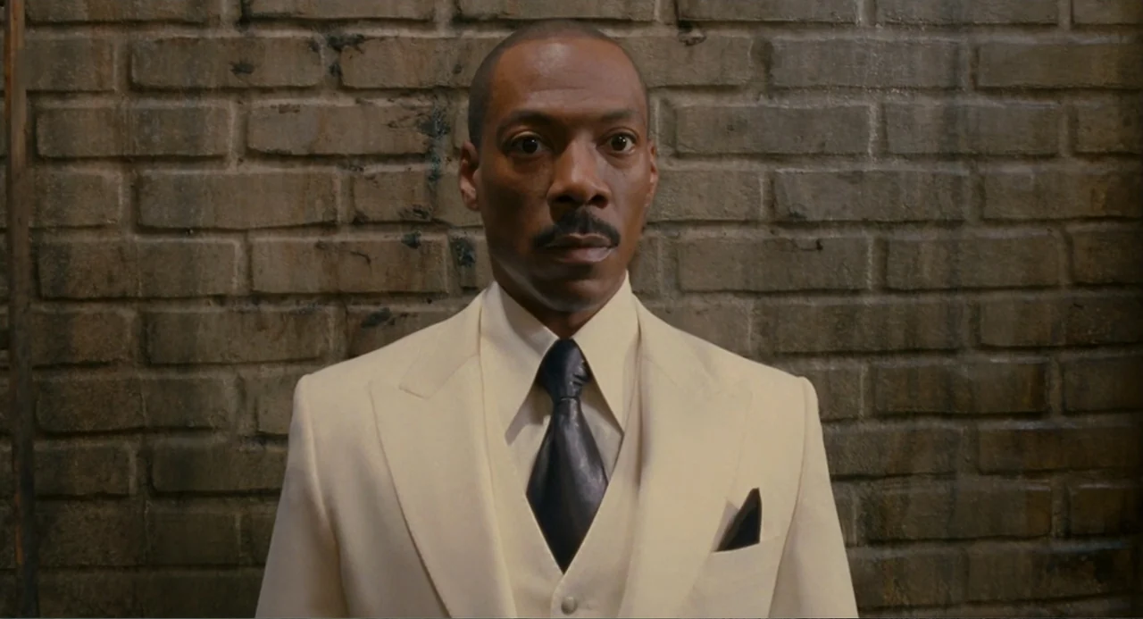 Meet Dave (2008) movie still: medium — A man stands centered in front of a weathered brown brick wall. The subject wears a formal cream suit while…