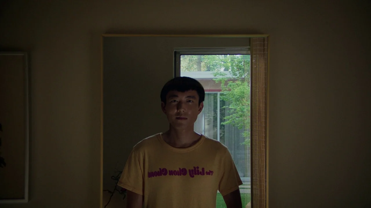 After Yang (2021) movie still: medium — A dimly lit room features a large mirror reflecting a window and outdoor greenery. A young man stands in…