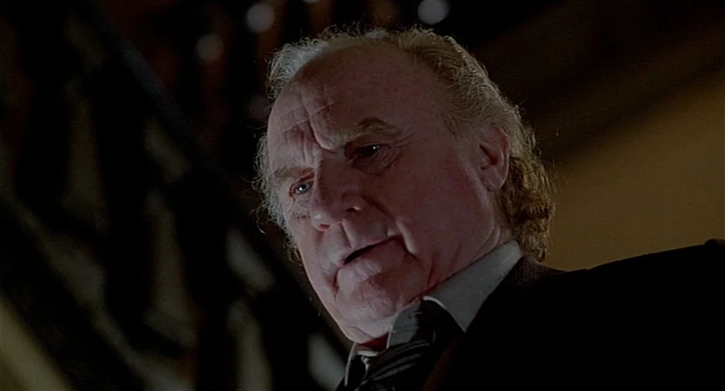 The Verdict (1982) movie still: closeup — An elderly man peers down from the top of a dark, shadowed wooden staircase. He stands alone in the dimly…