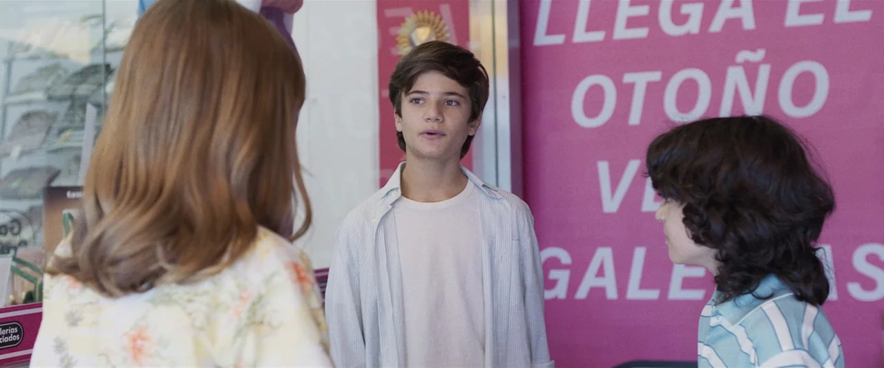 Voy a pasármelo bien (2022) movie still: medium — A brightly lit retail shop interior features a large magenta sign on the wall. Three young people…
