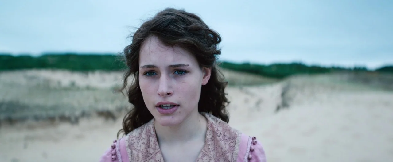 Slack Bay (2016) movie still: closeup — A sandy landscape with dunes under a flat, overcast sky. A young woman stands in the foreground, facing…