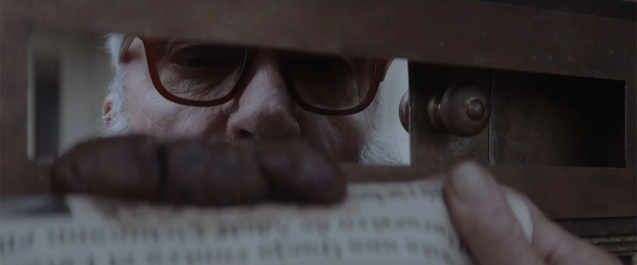 My Neighbor Adolf (2022) movie still: closeup — A man peers through a narrow horizontal mail slot cut into a wooden door. The man's eyes are visible…