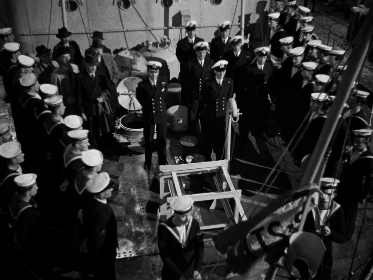 In Which We Serve (1942) movie still: wide — A crowded deck of a naval ship with officers and sailors in formation. The men stand in rows on a metal…