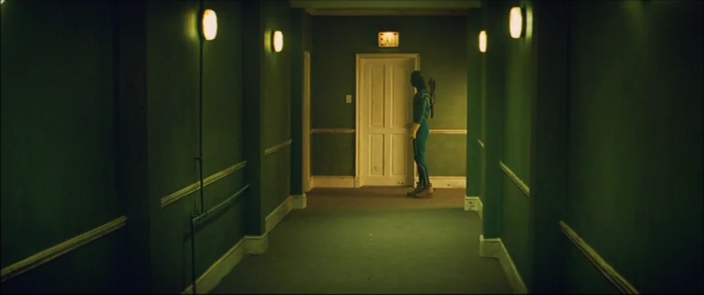 Kick Ass (2010) movie still: wide — A dimly lit narrow hotel hallway with green walls and mounted sconce lighting. A costumed figure stands alone in…