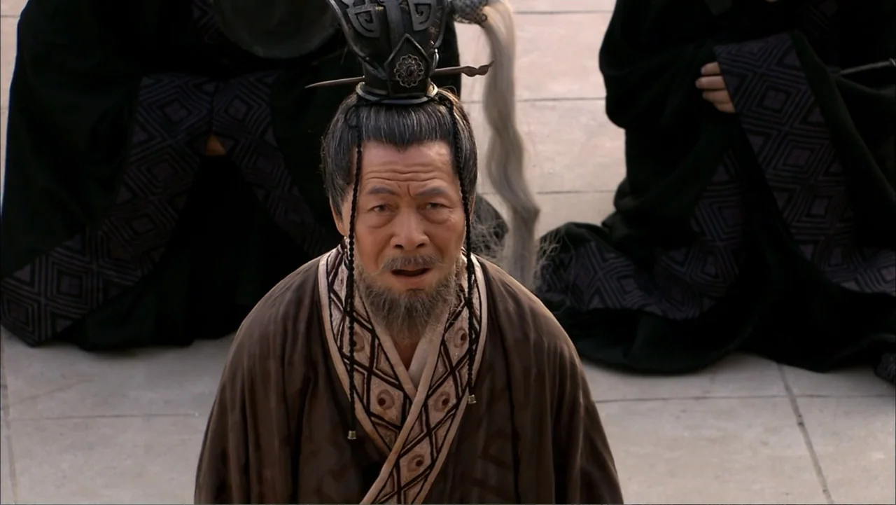 The First Emperor (2006) movie still: medium — A stone courtyard serves as the backdrop for several seated individuals in dark robes. A man with a…