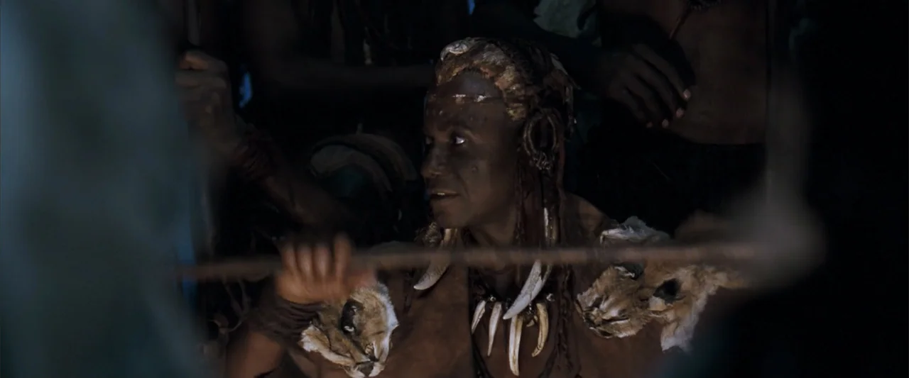 10,000 BC (2008) movie still: medium — A dark, cramped stone cave interior with shadows obscuring the surroundings. A man with tribal markings and…