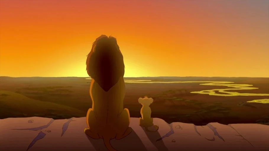 The Lion King (1994) movie still: wide — A vast savannah landscape extends below a rocky outcrop under a warm sunrise sky. An adult lion and a small…