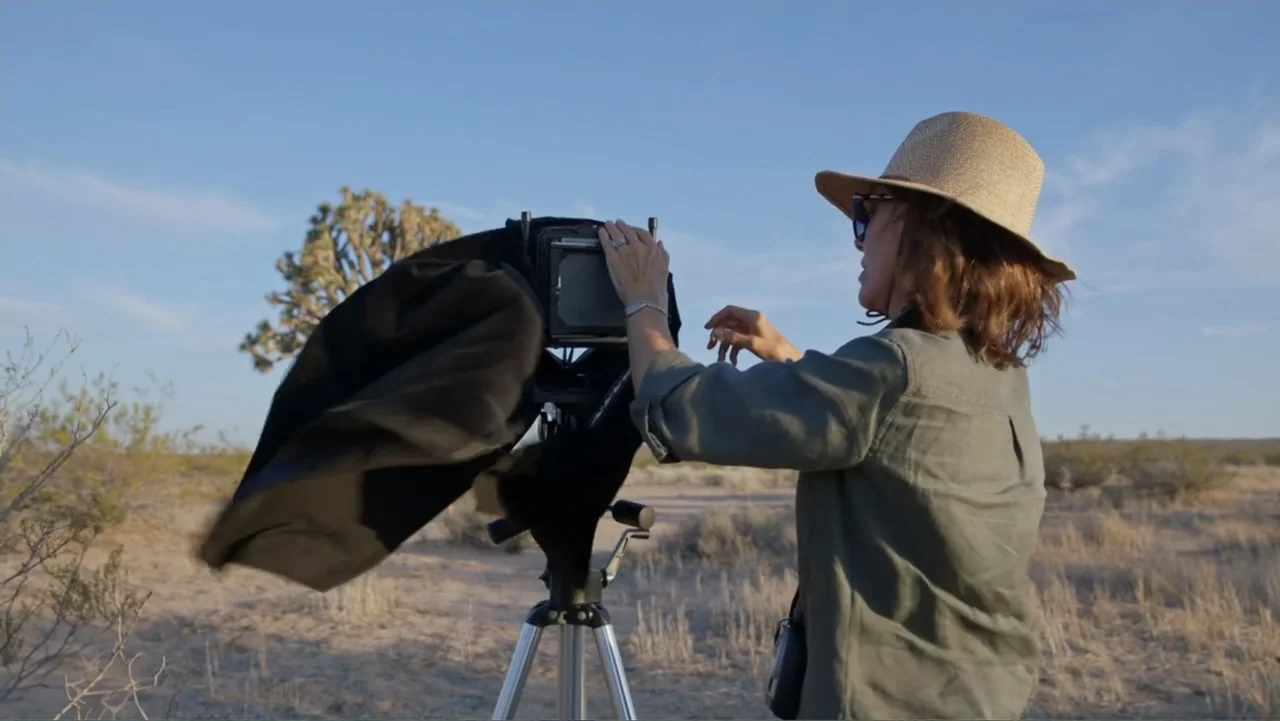 Trees and Other Entanglements (2023) movie still: medium — A woman stands in a dry desert landscape featuring sparse vegetation and a clear sky. She…