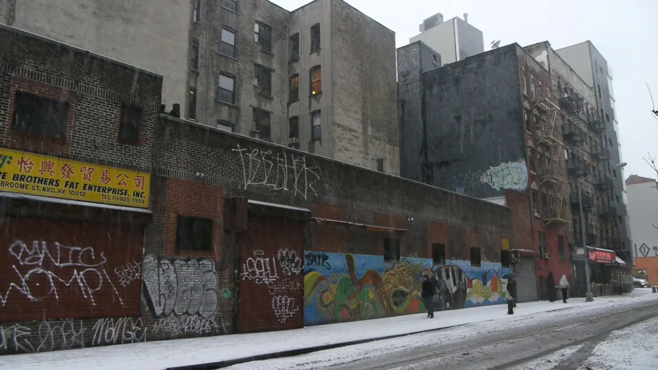 Counting (2015) movie still: wide — A snowy street in a dense urban neighborhood features brick buildings covered in graffiti. Pedestrians walk…