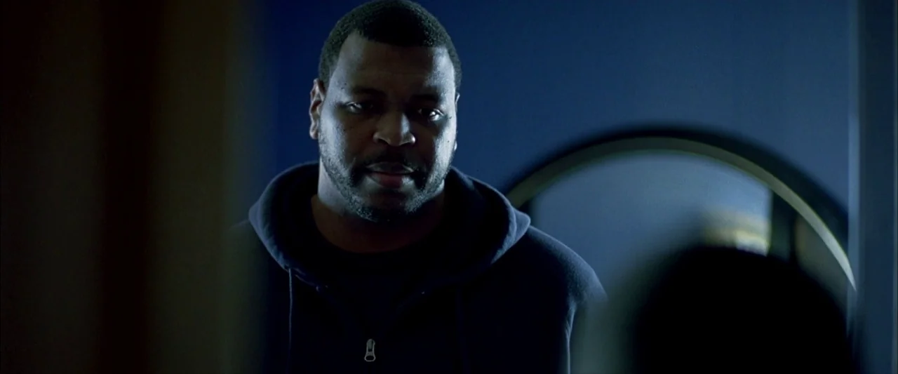Ballast (2008) movie still: closeup — A dark indoor space with a blue wall and a circular mirror. A man stands in the foreground looking forward.
