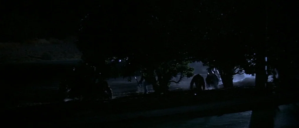 Assault on Precinct 13 (1976) movie still: wide — A dark, silhouetted line of trees obscures a patch of illuminated ground at night. Three…