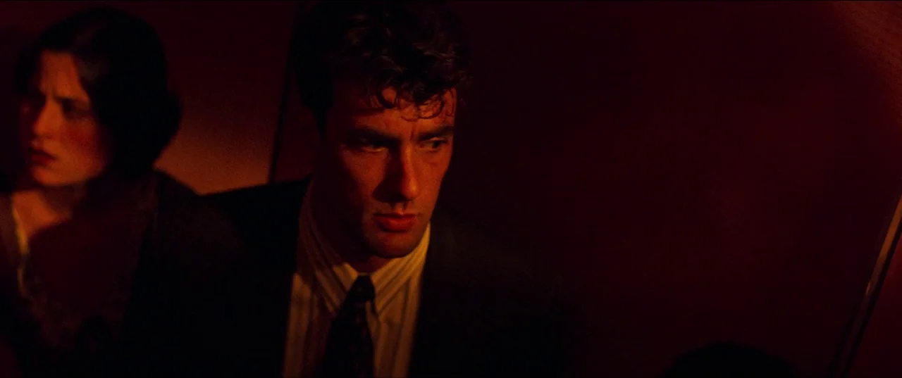 Speed (1994) movie still: medium — The interior of a darkened train carriage features deep red walls and low lighting. A man in a suit looks away…