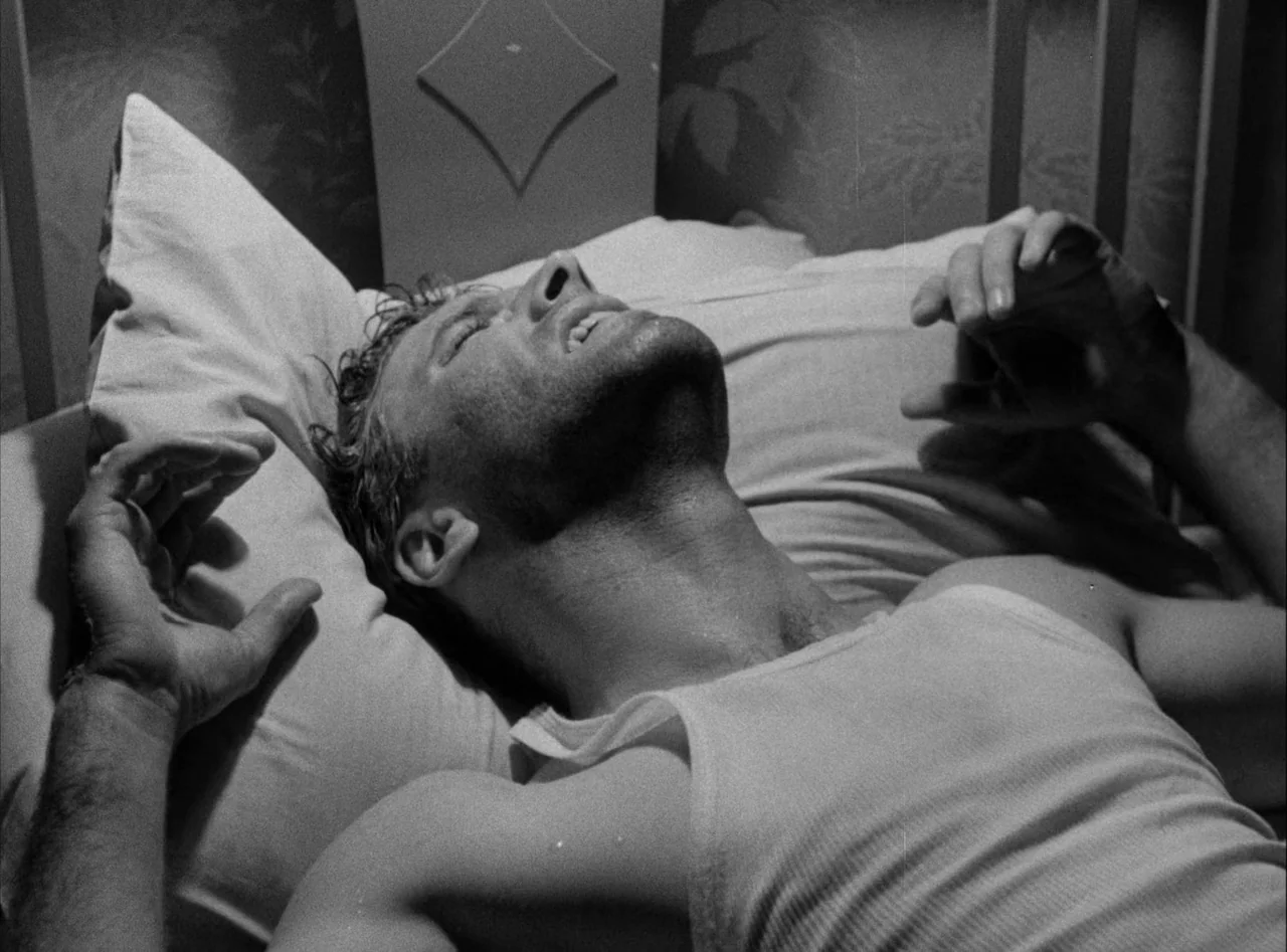 The Killers (1946) movie still: closeup — A man lies on a pillow in a dimly lit, high-contrast room. He rests with his eyes closed and hands raised…