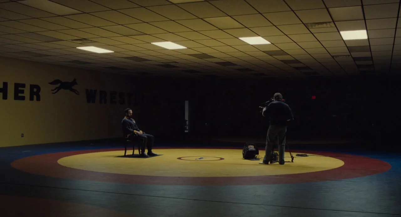 Foxcatcher (2014) movie still: wide — A vast, dark interior wrestling training facility with a circular mat in the center. A man sits on a chair in…