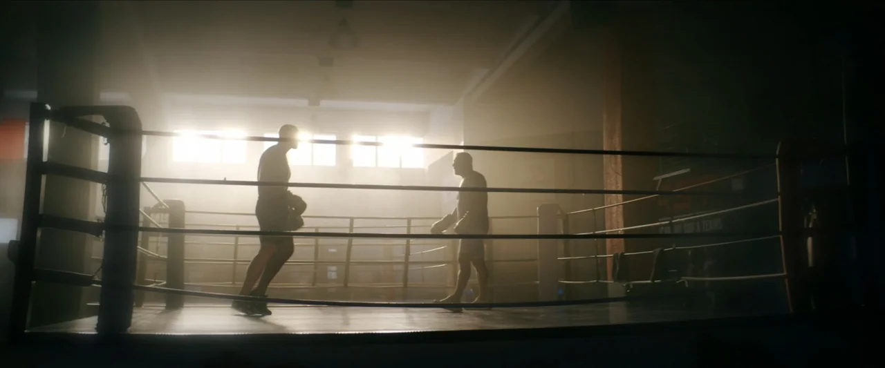 Black Lotus (2023) movie still: wide — A dimly lit gym interior features a raised boxing ring with ropes and haze. Two athletes in athletic gear…