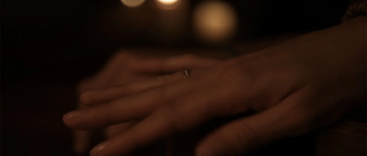 La coherencia del caos movie still: closeup — A dark indoor space is illuminated by soft light in the background. A human hand rests on a dark…