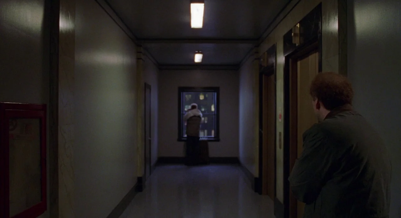 Adaptation (2002) movie still: wide — A long apartment building hallway is dimly illuminated by overhead lights and a single window. A man in the…