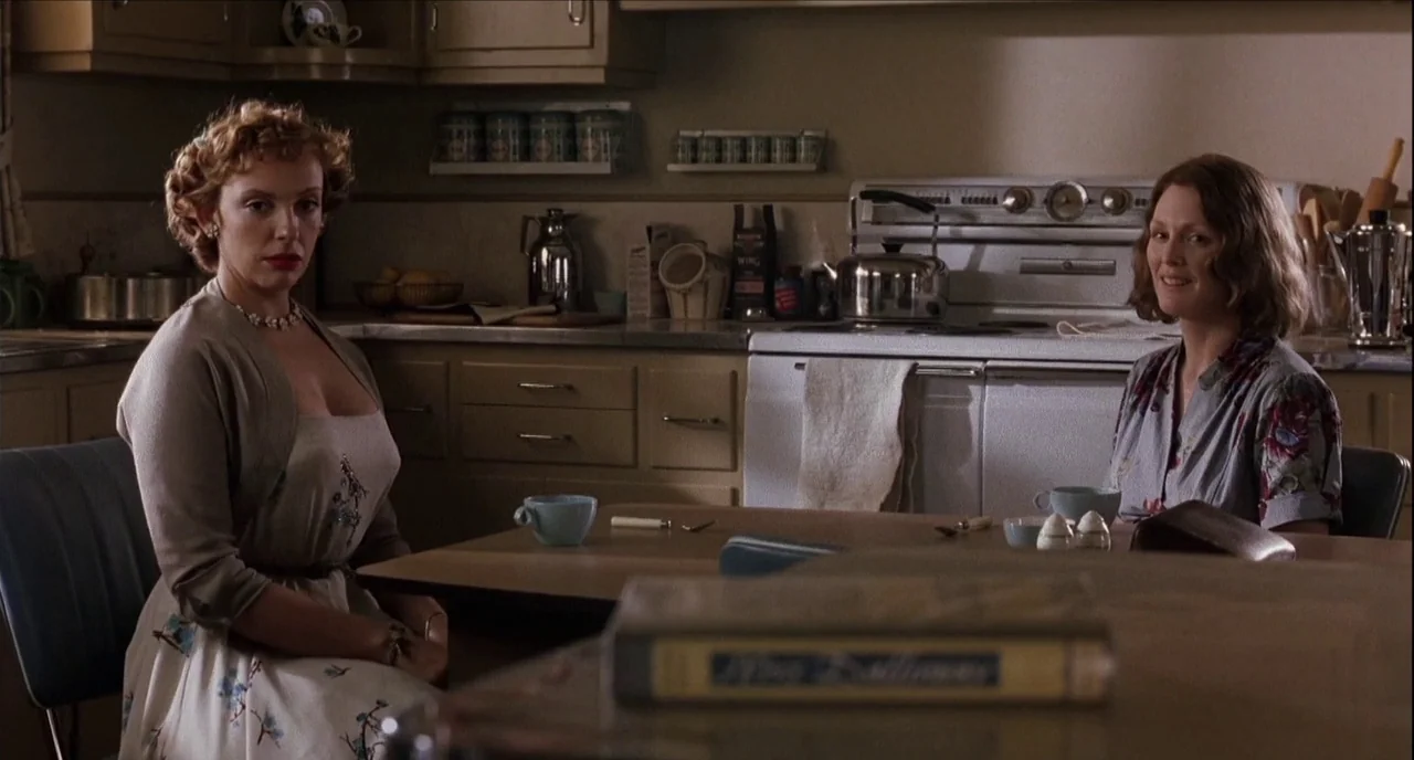 The Hours (2002) movie still: medium — A 1950s-style kitchen features tan wooden cabinets and a vintage white range stove. Two women sit at a table…