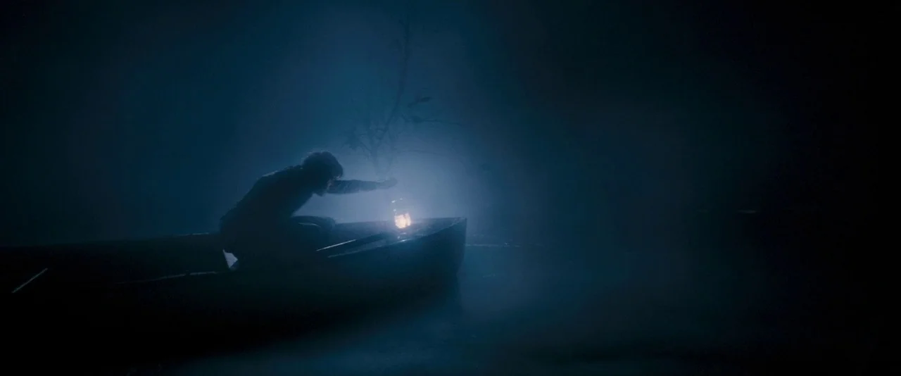 Dead Silence (2007) movie still: wide — A small boat drifts on a dark, foggy body of water at night. A person kneels in the boat, holding a glowing…