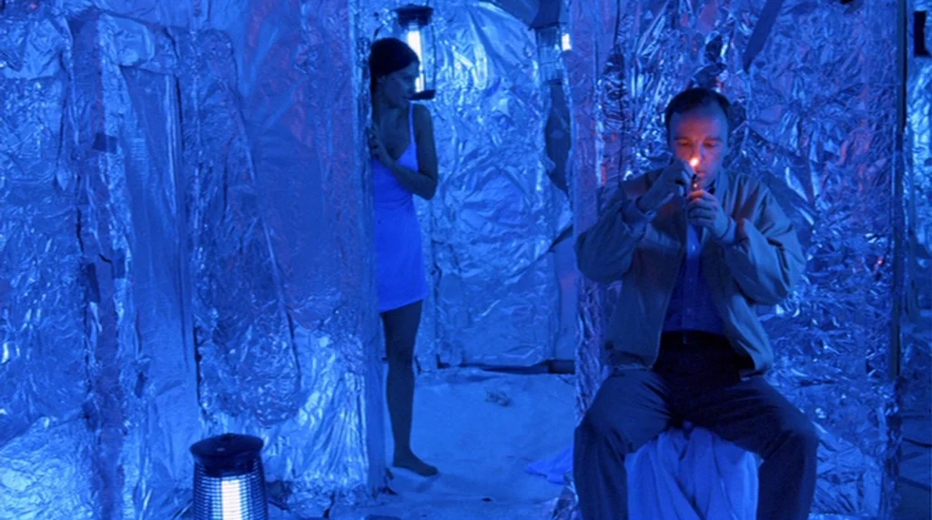 Bug movie still: medium — A small interior space covered entirely in crinkled aluminum foil with intense blue lighting. A man sits on a low stool…