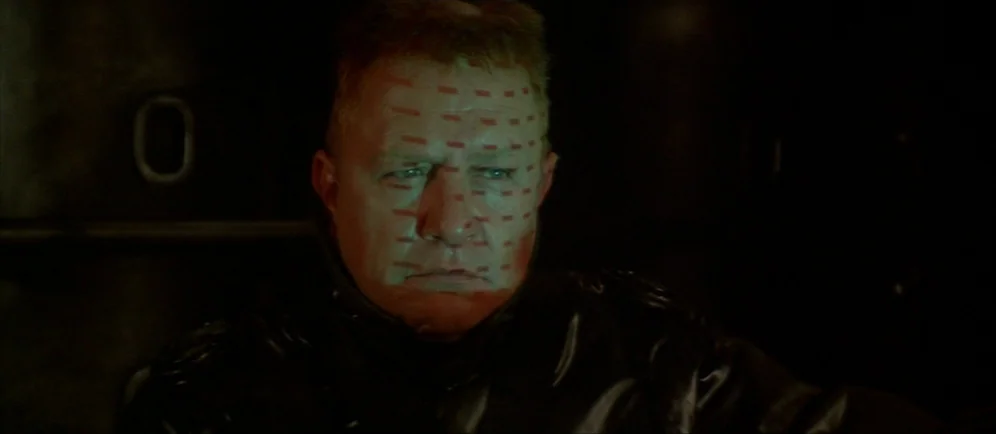 Dune (1984) movie still: closeup — A dark vehicle interior with metallic surfaces surrounding the subject in low light. A red laser grid is…