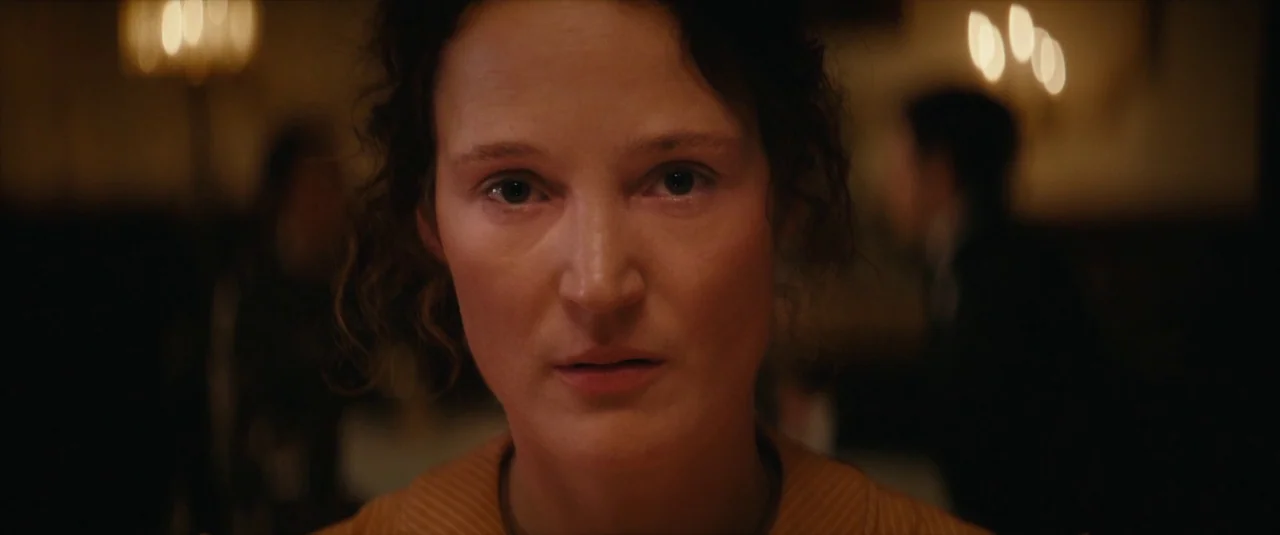 The Dead Don’t Hurt (2023) movie still: closeup — A dimly lit indoor dining area features soft out-of-focus lights in the background. A woman in a…