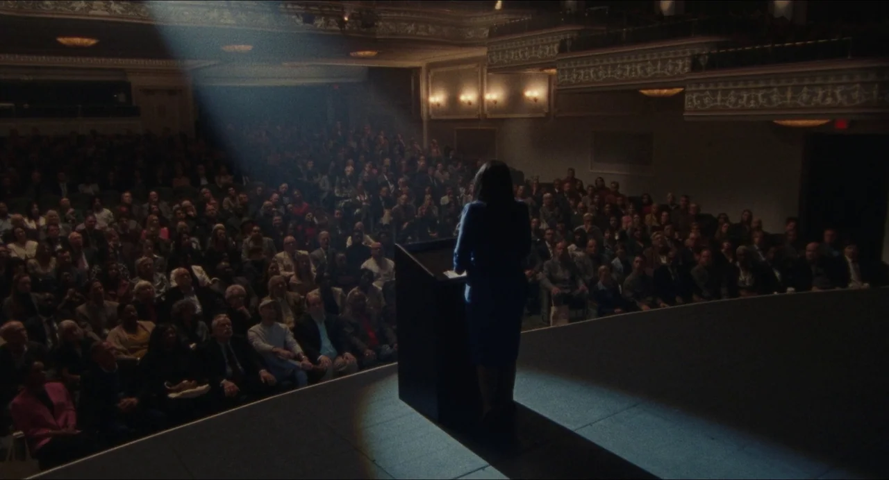 Origin (2023) movie still: wide — A dark, ornate theater auditorium filled with a large crowd facing a central podium. A woman in a dark suit stands…