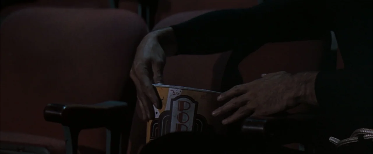 The Pelican Brief (1993) movie still: closeup — A dark movie theater interior with red fabric seats and a paper popcorn container. A person sits in…