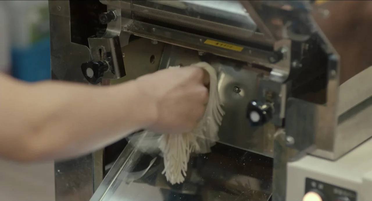 Ramen Shop (2018) movie still: closeup — A stainless steel noodle machine sits on a counter in a sterile food preparation area. A person's hand…