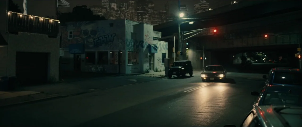 Guns Up (2025) movie still: wide — A quiet urban street at night features graffiti-covered low-rise buildings and an overhead bridge. A lone car…
