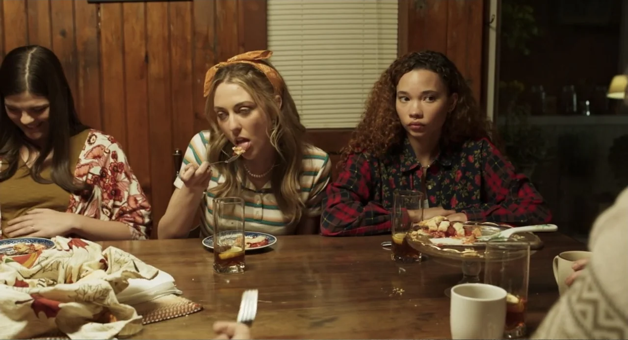 Salvation (2025) movie still: medium — Three young women sit around a wooden dining table in a wood-paneled room. They eat pie and drink iced tea…