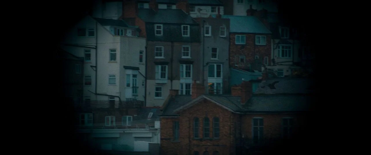 Saint Maud (2019) movie still: establishing — Stacked residential buildings are arranged on a hillside under a dark, nighttime sky. Dark brick and…