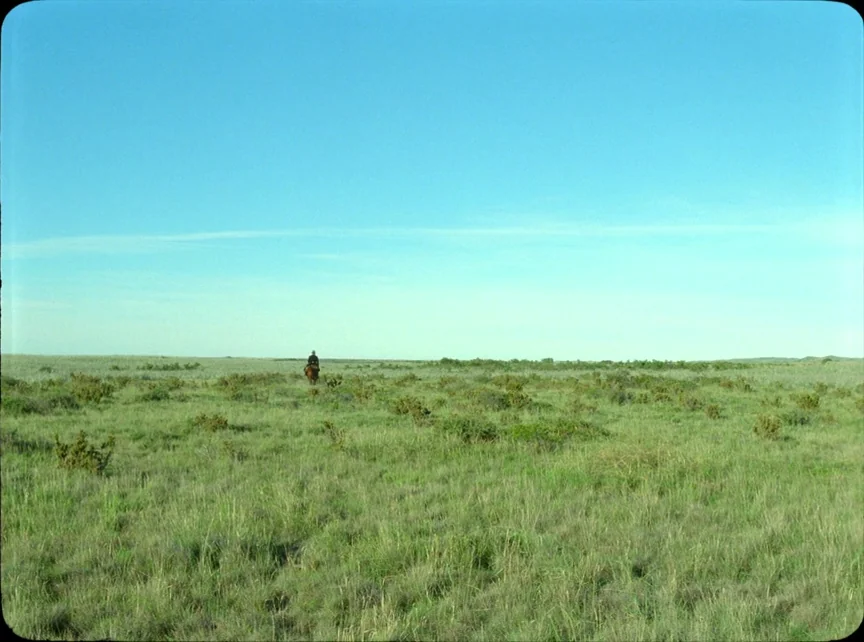 Jauja (2014) movie still: wide — A vast, flat expanse of green grassland stretches under a clear blue sky. A lone rider on horseback slowly moves…