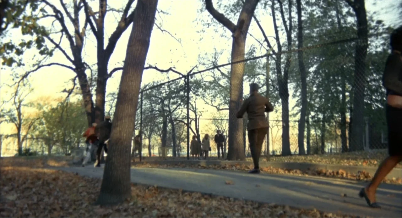 Cooley High (1975) movie still: wide — A park pathway is bordered by autumn trees and a high chain-link fence. Several individuals walk along the…