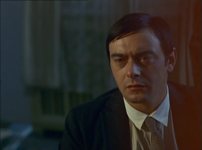 Satan’s Brew (1976) movie still: closeup — A dimly lit room with draped curtains visible in the background out of focus. A man with dark hair in a…