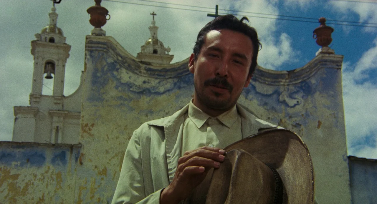 Canoa: A Shameful Memory (1976) movie still: medium — An aged, weathered church with blue and yellow stucco sits against a cloudy sky. A man stands…