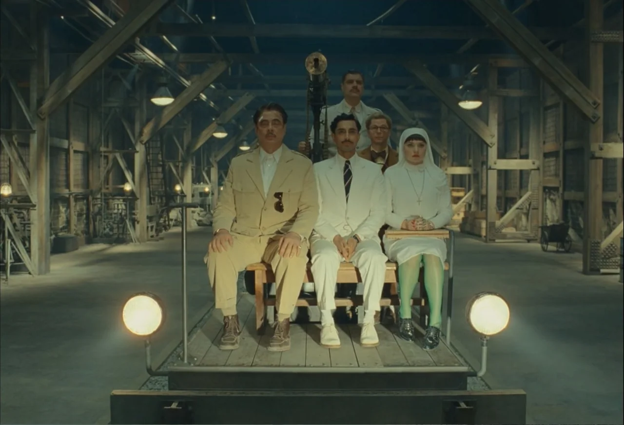 The Phoenician Scheme (2025) movie still: fullbody — A spacious wooden workshop features complex support beams and dark industrial flooring. Four…