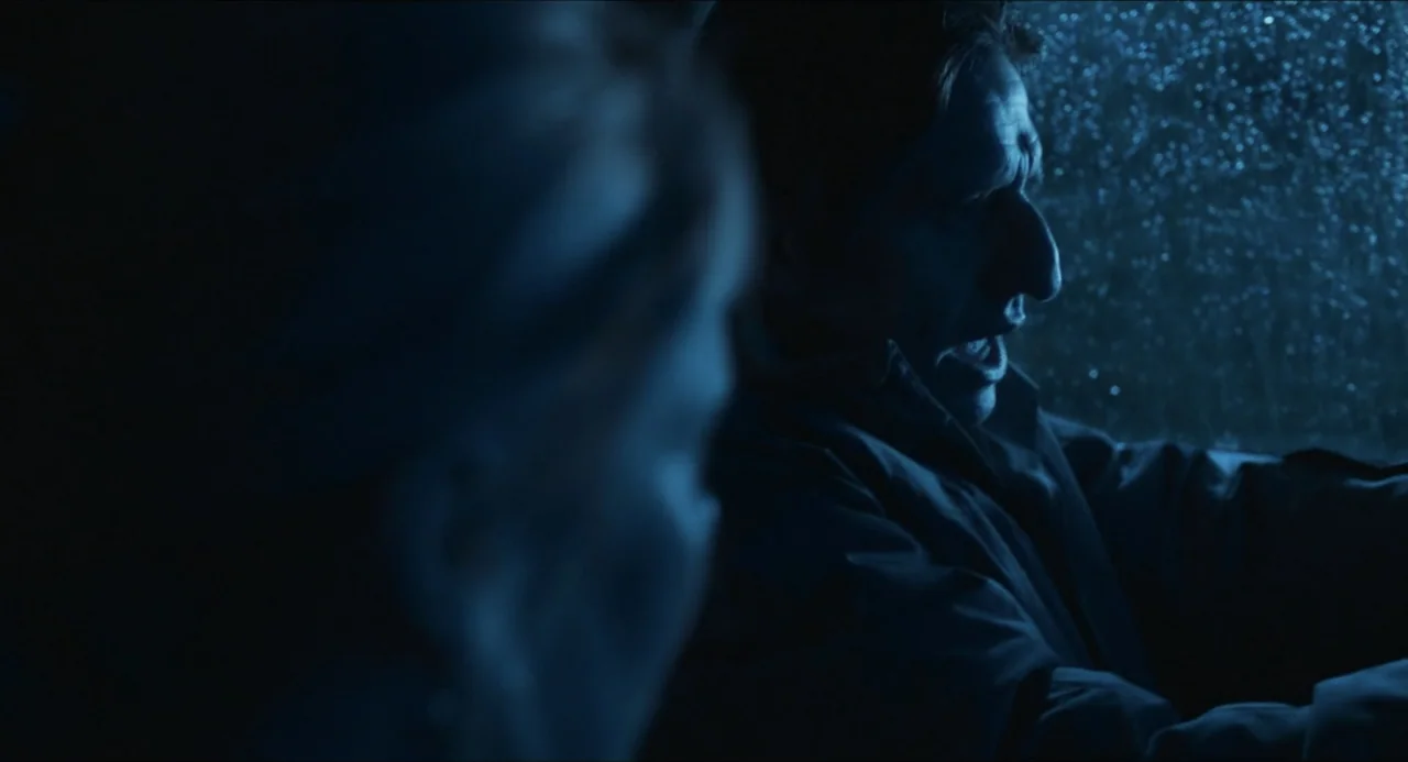 Echoes Of A Crime (2025) movie still: medium — The interior of a car at night with rain visible on the window glass. A man sits in the driver seat…