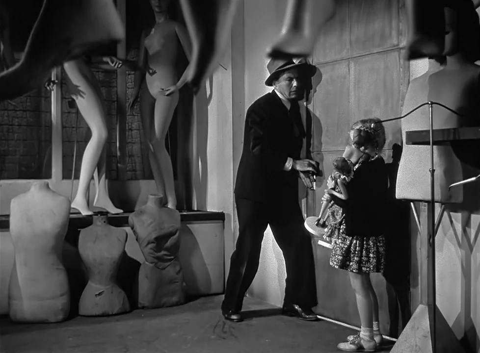 M (1951) (1951) movie still: medium — A dark, cramped room is crowded with various plaster mannequins and mannequin limbs. A man in a suit…