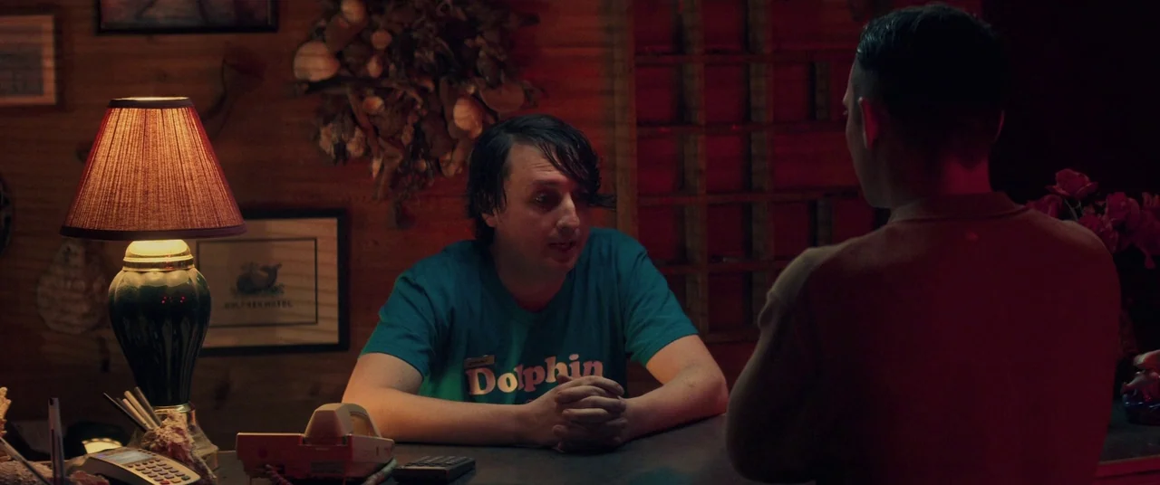Come To Daddy (2019) movie still: medium — A dimly lit hotel reception area features wood-paneled walls decorated with shell art. A man behind the…