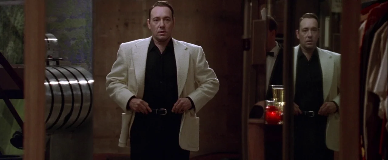 L.A. Confidential (1997) movie still: medium — A dimly lit room with wooden wall paneling and a large vertical mirror present. A man stands before…