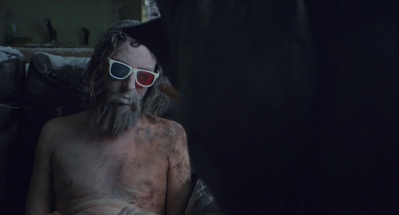 Relaxer (2018) movie still: medium — A dimly lit, dusty indoor space provides the setting for the interaction. A man covered in dust wears 3D…