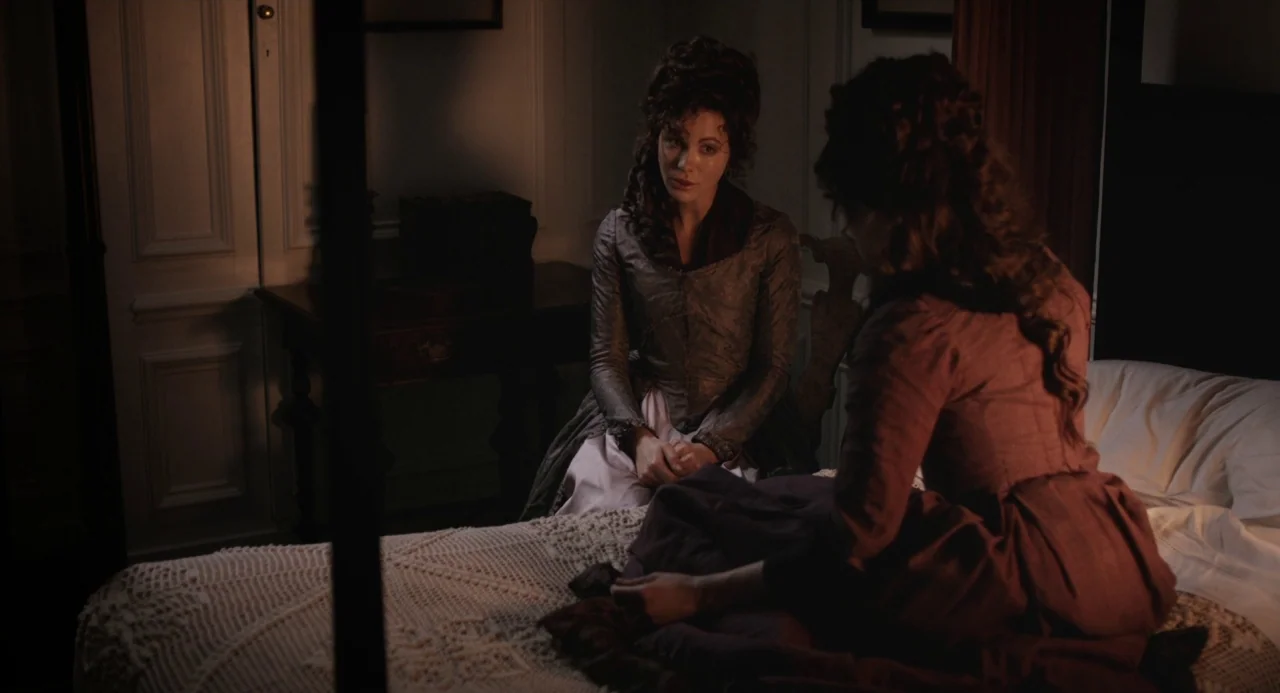 Love and Friendship (2016) movie still: medium — A dimly lit historical interior features two women sitting on and near a bed. One woman sits on the…