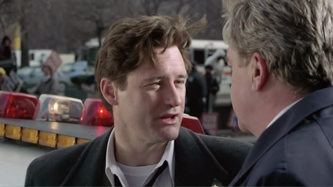 Mistrial (1996) movie still: medium — An outdoor urban location with a police vehicle parked in the background. A man with brown hair speaks to an…