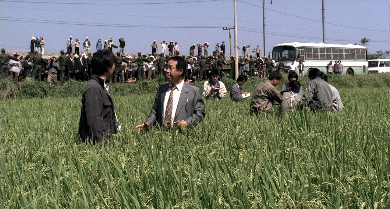 Memories of Murder (2003) movie still: wide — A crowd of people gathers behind a wire fence near a large white bus. Two men in suits stand in the…